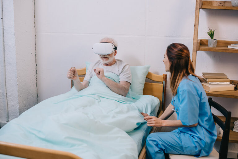 personne âgée avec un casque de réalité virtuelle dans son lit, avec à son chevet une aide soignante